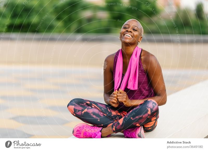Cheerful back sportswoman with towel sitting on embankment after workout break activewear healthy pavement city building relax wellness fit sportswear
