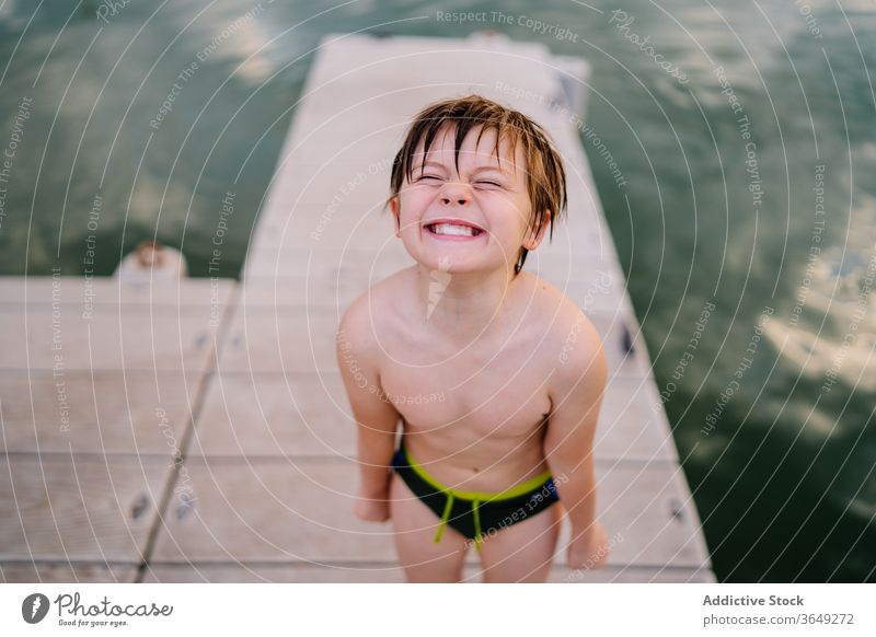 Delighted boy on pier near lake make face smile cheerful swimwear quay funny grimace happy joy kid child childhood pond water summer rest relax cute having fun