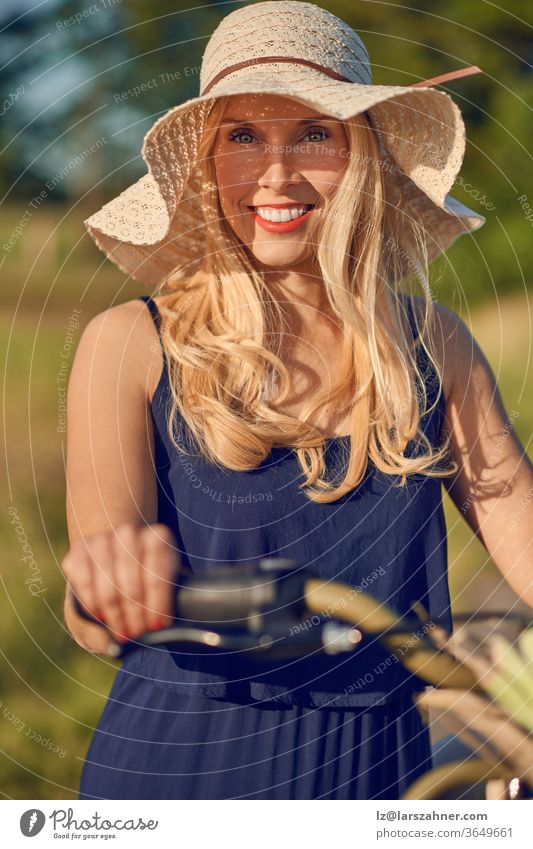 Attractive blond woman with lovely friendly smile standing outdoors with her bicycle in the spring sunshine wearing a broad brimmed straw sunhat attractive
