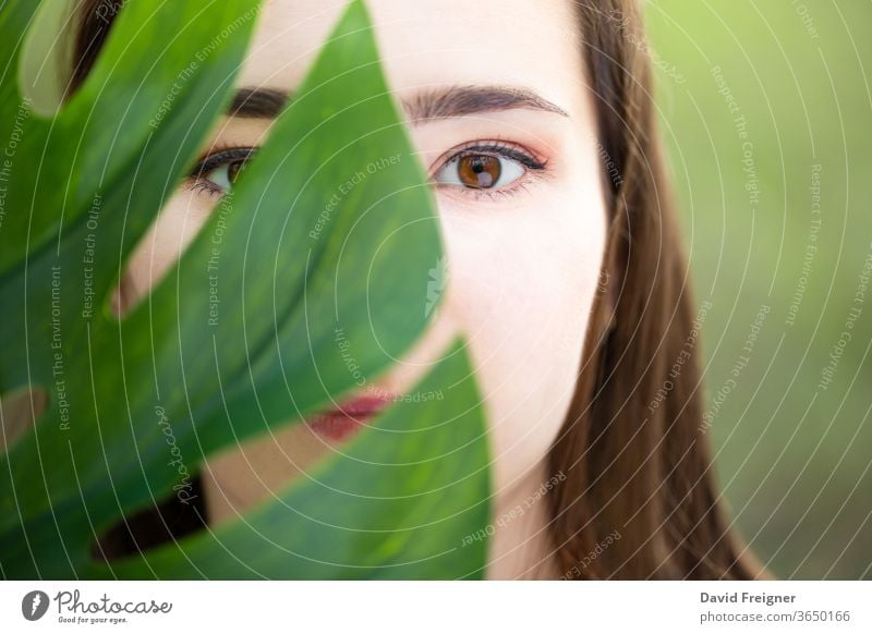 Beautiful natural young woman closeup behind big monstera leaf with green background in the woods. skin beautiful girl beauty health model female nature