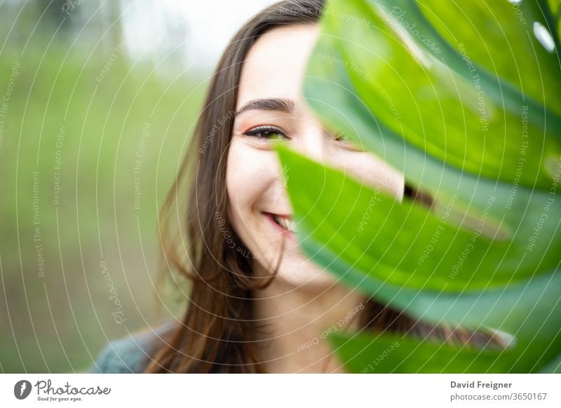 Beautiful natural young woman closeup behind big monstera leaf with green background in the woods. smile face attractive clean cute organic purity pure eyes