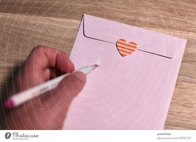 Left-handed. One hand holds a pen in writing position. An envelope closed with a heart sticker is addressed. Love letter. Mail enveloped in a letter