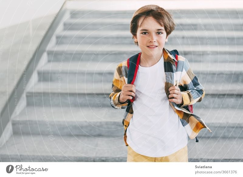 A happy boy with a backpack stands on the steps in front of the entrance to the school and smiles beautifully. Beginning of the new school year after the summer holidays. Back to school