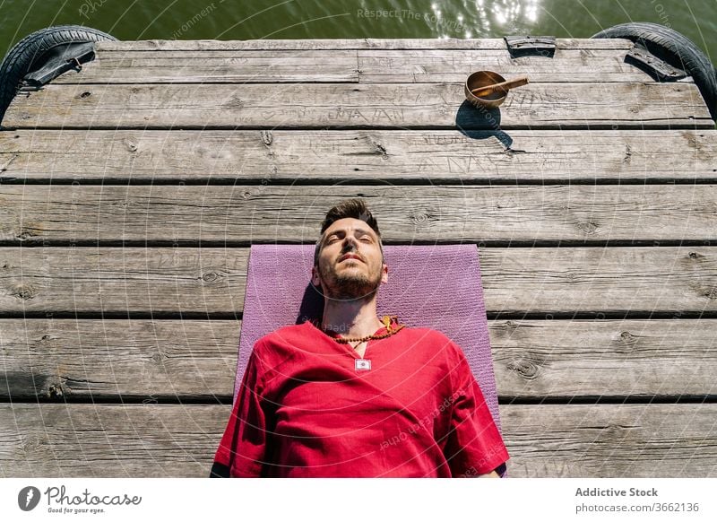 Relaxed man meditating on wooden pier near singing bowl after yoga training lying mat meditate relax embankment recreation eyes closed seaside session male
