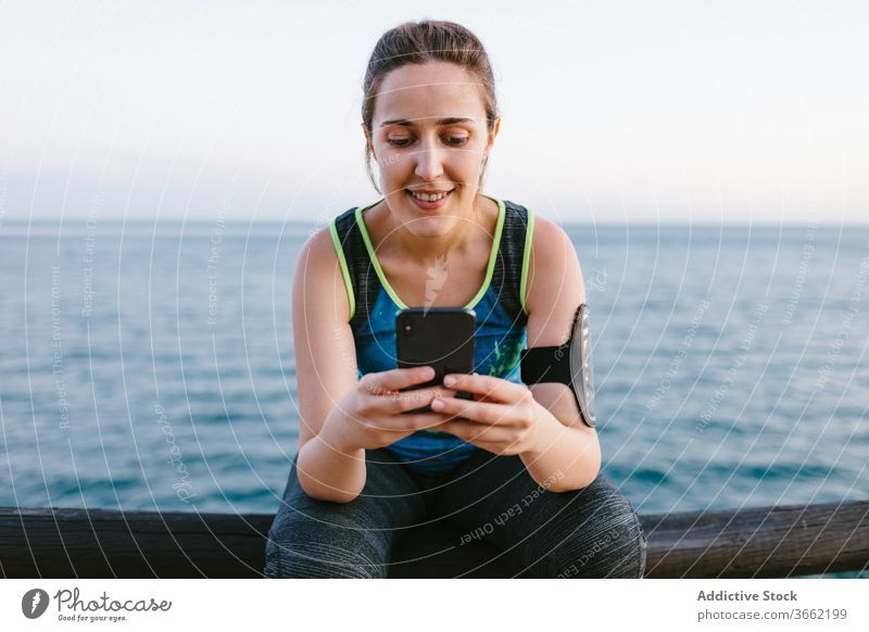 Smiling woman in sportswear using smartphone near sea sportswoman embankment browsing relax training smile female fitness cellphone workout device gadget sporty