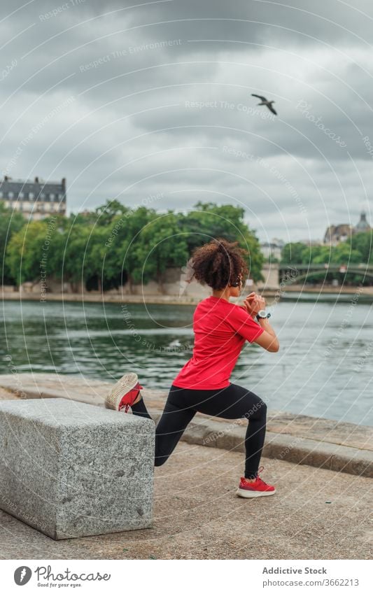 Unrecognizable sportswoman stretching legs near city river during workout exercise activewear embankment smart watch device overcast weather urban pond training