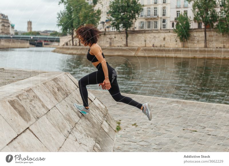 Unrecognizable ethnic sportswoman working out on embankment near city river workout warm up training climb platform cardio pond energy athlete reached leg