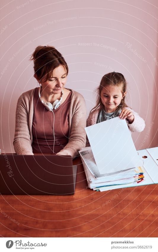 Woman working talking doing her job remotely during video chat phone call on laptop from home. Woman sitting at desk in front of computer looking at screen using headphones and smartphone while her daughter playing around. Concept of remote working