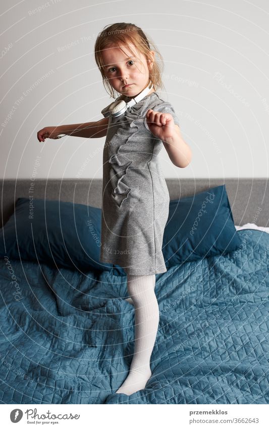 Little girl singing holding headphones cord imitating herself a real singer. Child having fun jumping dancing listening to music on bed in bedroom at home child