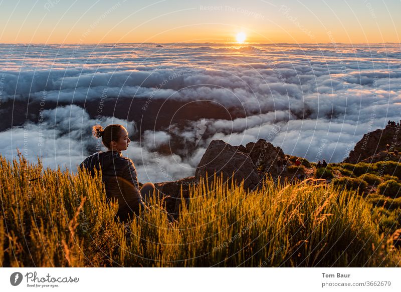young woman at the edge of a mountain peak sits above the clouds and watches the sunrise. The rising sun on the horizon close above a dense carpet of clouds