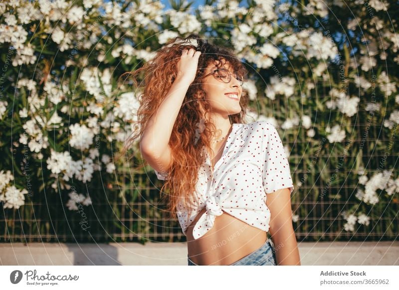 Positive female with wavy hair standing near blooming flowers woman touch hair fence happy beauty lifestyle belly idyllic feminine summertime toothy smile