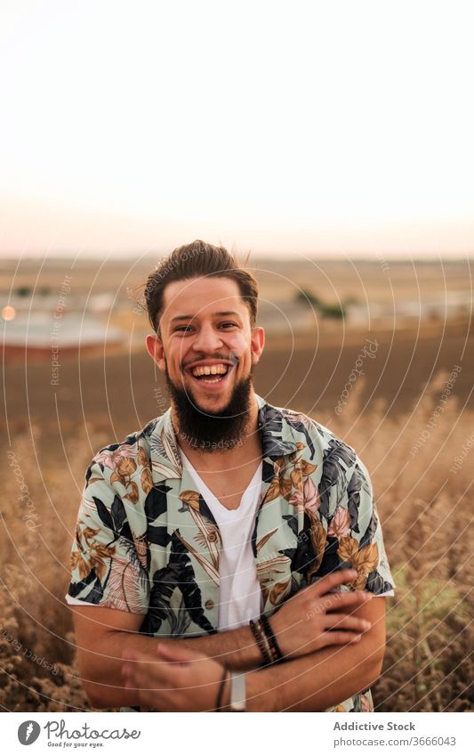 Happy hipster man laughing in field at colorful sunset enjoy slim grass countryside lifestyle positive pleasure evening confident toothy smile vacation