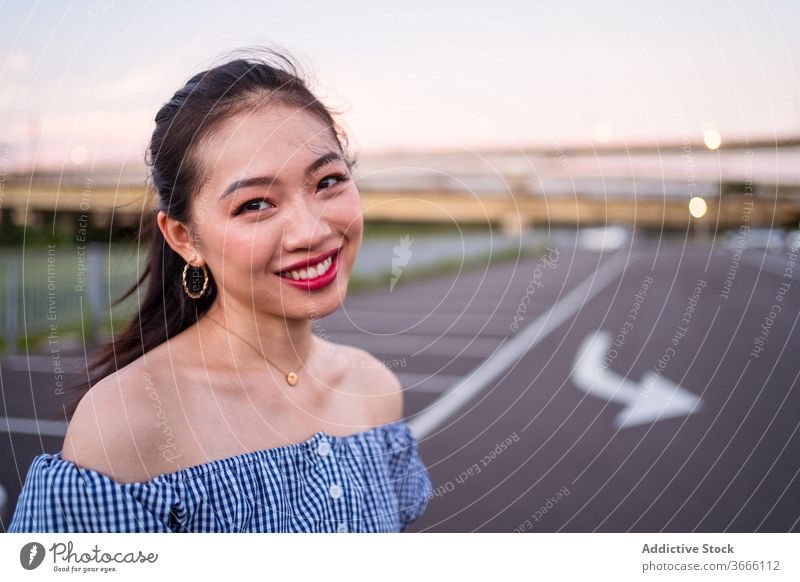 Sincere smiling Asian woman standing on city road in summer sincere accessory smile marking calm sky tranquil satisfied quiet golden earring necklace asphalt