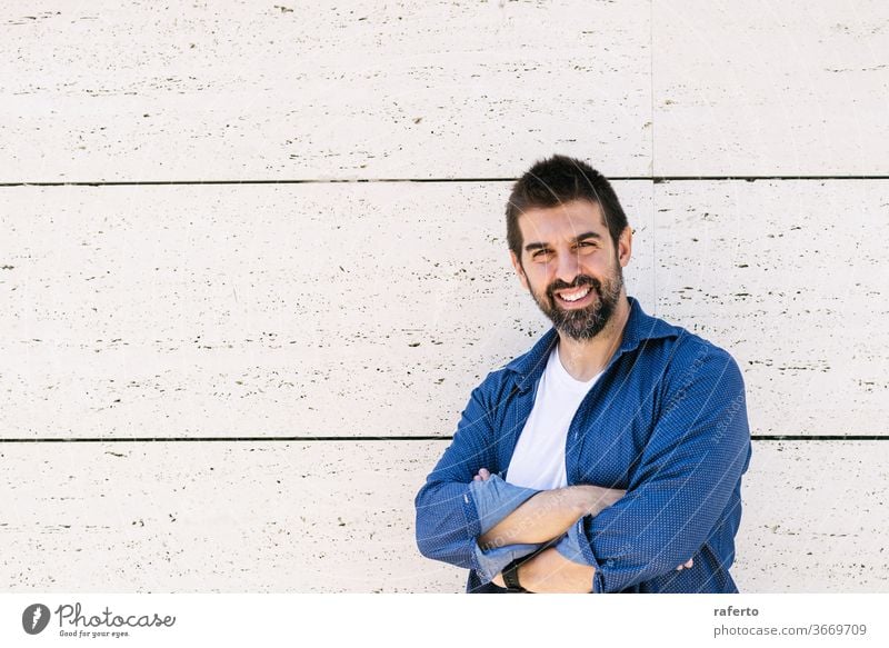 Bearded man leaning on wall with crossed arms while looking camera with a smile 1 male standing bearded cheerful attractive hispanic arms crossed casual attire