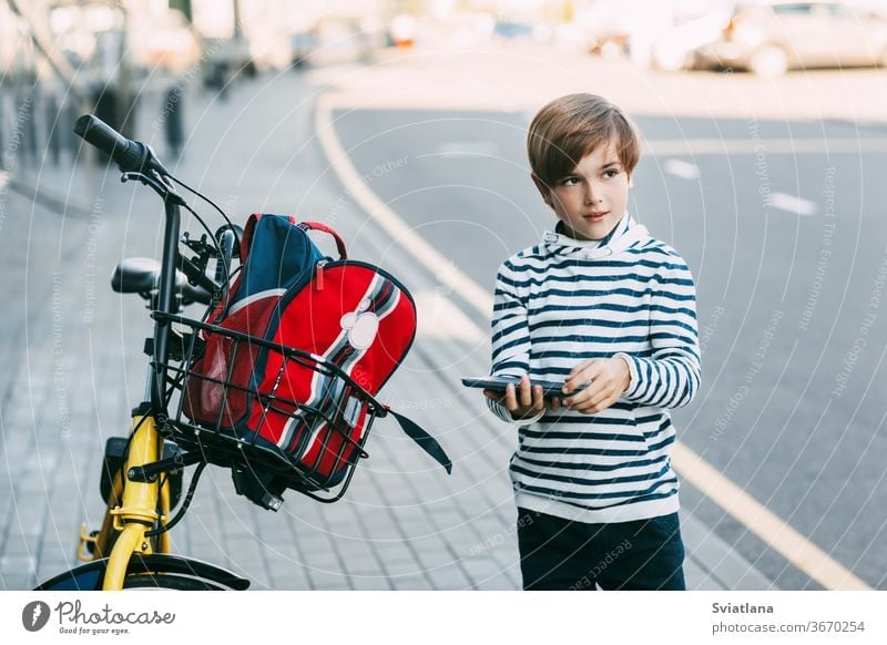 A cute boy in a striped sweater holds a tablet in his hands and stands next to a Bicycle with a backpack hanging from it. The boy is going to ride home after school on a Bicycle. Safe way home