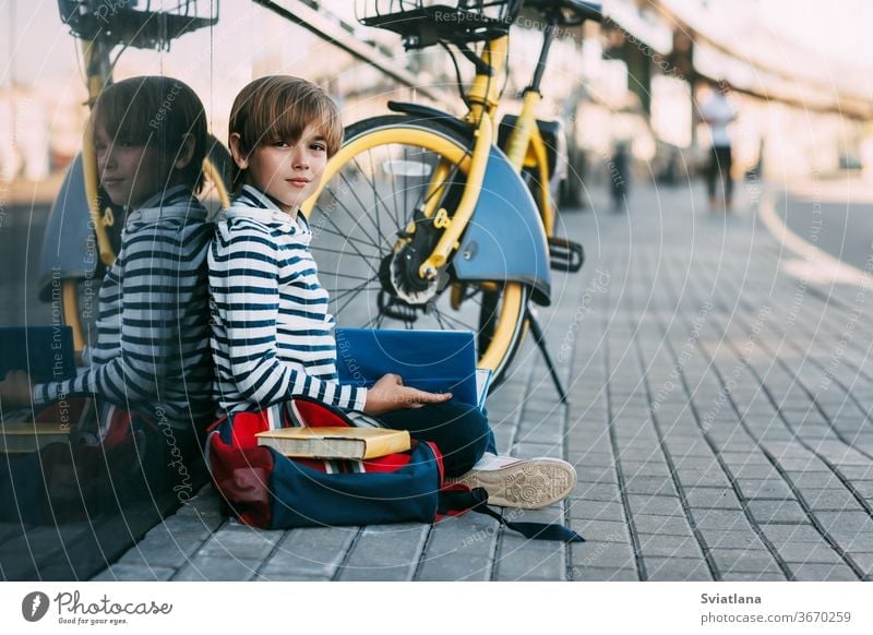 A cute schoolboy is sitting in the open air at school and holding a book in his hands. Next to it is a school backpack and a Bicycle, space for text face tired