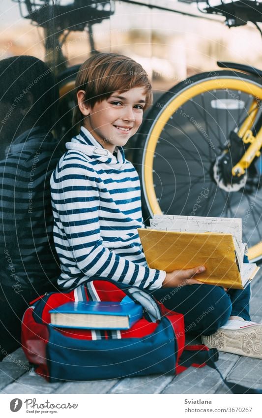 A cheerful boy is sitting on the sidewalk near the school and reading a book. Next to it is a Bicycle and a backpack face tired cover education portrait holding