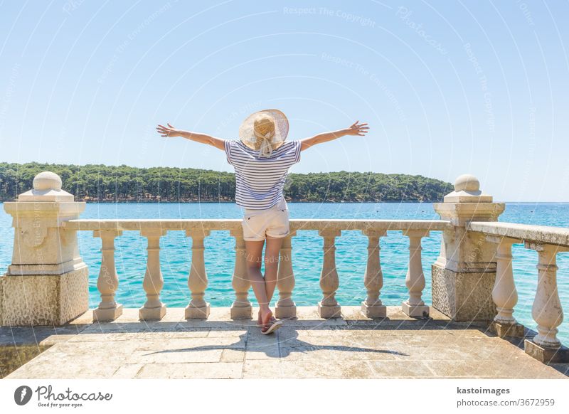 Rear view of happy woman on vacation, wearing straw summer hat ,standing on luxury elegant old stone balcony of coastal villa, relaxing, arms rised to the sun, looking at blue Adriatic sea.