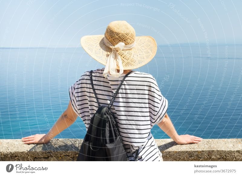 Rear view of a traveller wearing a straw hat and rucksack, leaning against a stone wall and looking out over the big blue sea and the islands on the horizon. Copy space.