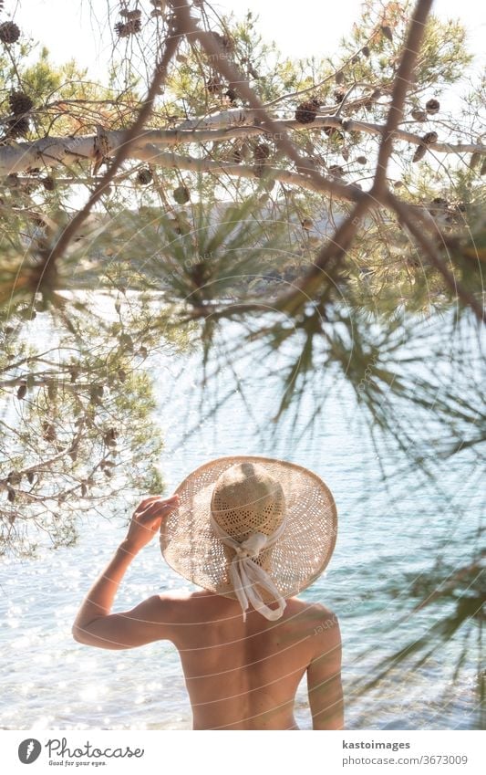 Rear view of topless beautiful woman wearing nothing but straw sun hat realaxing on wild coast of Adriatic sea on a beach in shade of pine tree. Relaxed healthy lifestyle concept.