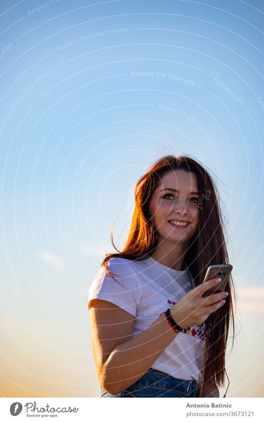Attractive brunette holding a smartphone laughing , beautiful sunny day. Sunset golden hour, hair glowing orange from the sun, clean blue sky 20s adult