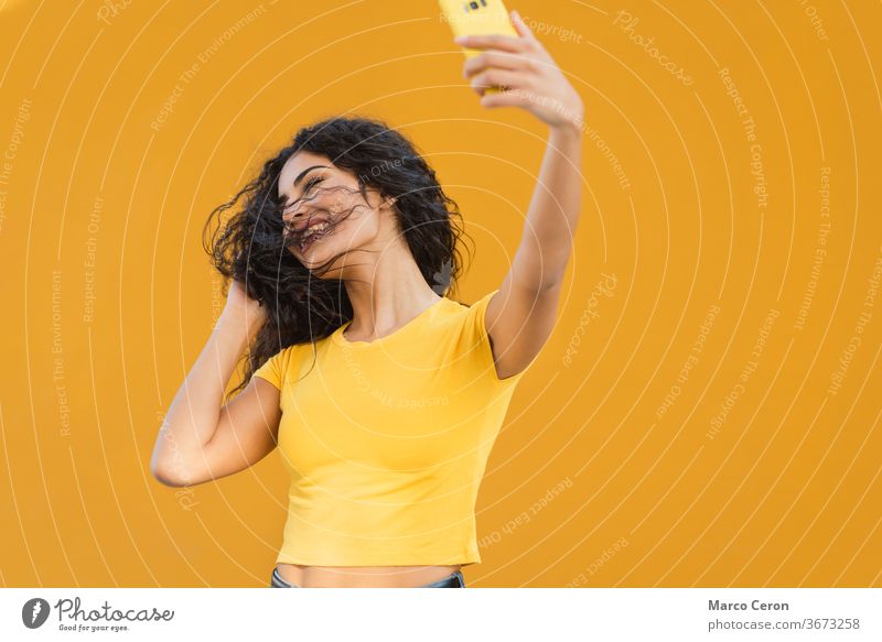 young mixed race woman inyellow t-shirt making a funny selfie with her phone with orange wall in the background taking smartphone lifestyle laisure activity