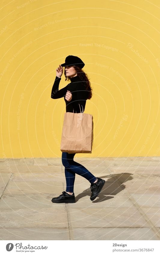 Fashion portrait woman with shopping bags wearing black outfit with hat posing on yellow wall. female girl sale happy beautiful adult fashion shopaholic young