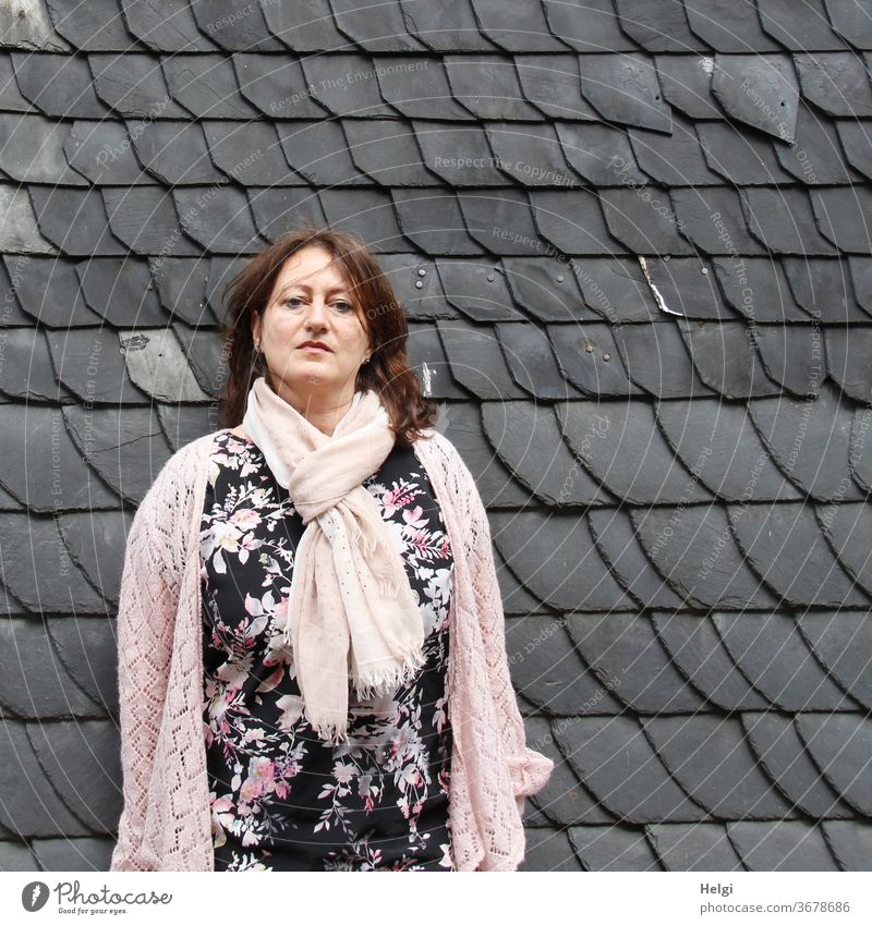 long-haired woman in black-pink-white flowered dress and pink jacket and scarf is standing in front of a grey shingle wall Woman Human being feminine Feminine