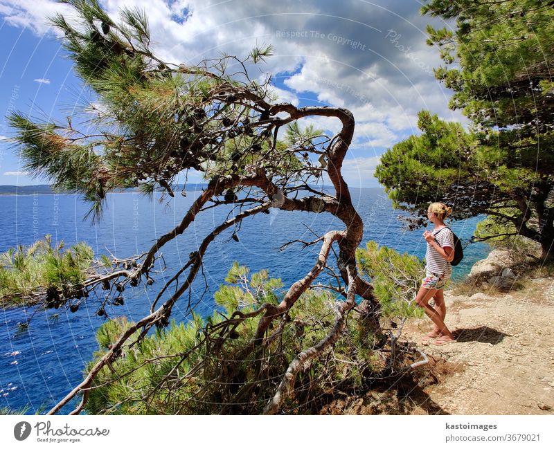 Young active feamle tourist wearing small backpack walking on coastal path among pine trees looking for remote cove to swim alone in peace on seaside in Croatia. Travel and adventure concept.