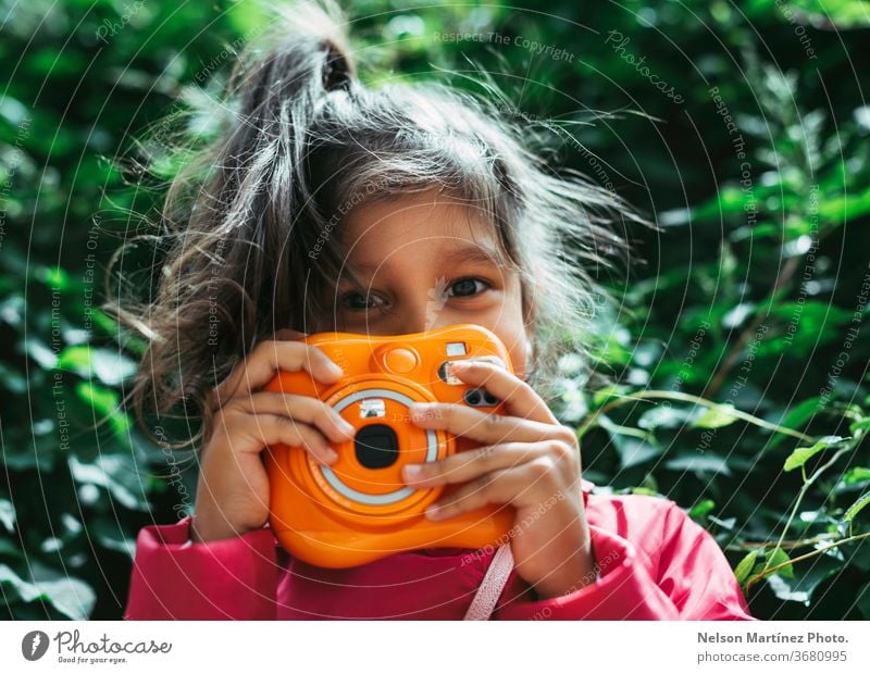 Little hispanic girl holding an Instax orange camera. Vintage camera. She is in front of a green background. kid little girl photography leasure fun colorful