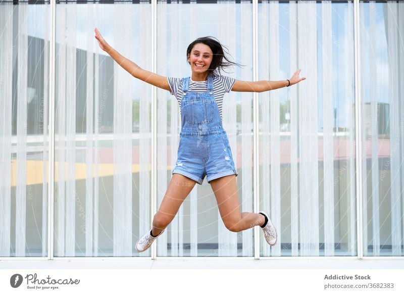 Happy teen girl jumping near glass wall smile happy casual leap fun excited building joy glad optimist modern delight teenage positive moment millennial