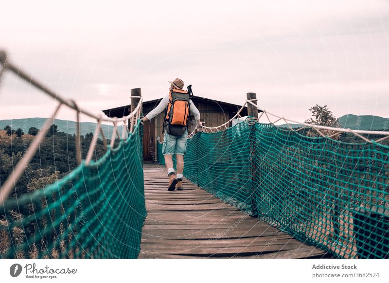 Anonymous traveling man walking along wooden footbridge suspension tourist backpack admire landscape vacation male adventure trip tourism content journey
