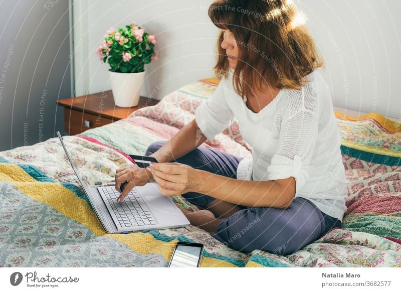 Woman paying by card for shopping online from home on a laptop, sitting on the bed. woman buy purchase middle aged activity bedroom business care caucasian