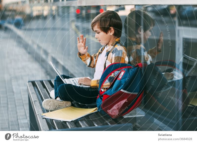 A cute boy is sitting on a bench, holding a laptop on his lap, next to a backpack. The boy communicates with friends using the computer, waving to them. Education, technology, distance learning