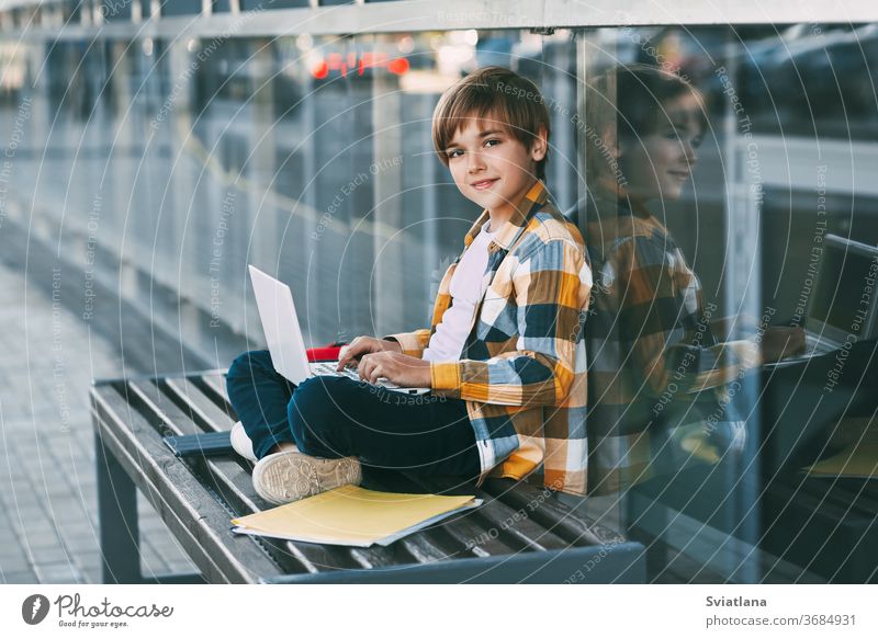 A cute boy in a plaid shirt is sitting on a bench with a laptop and typing on the keyboard, next to a backpack. The student is preparing for outdoor classes