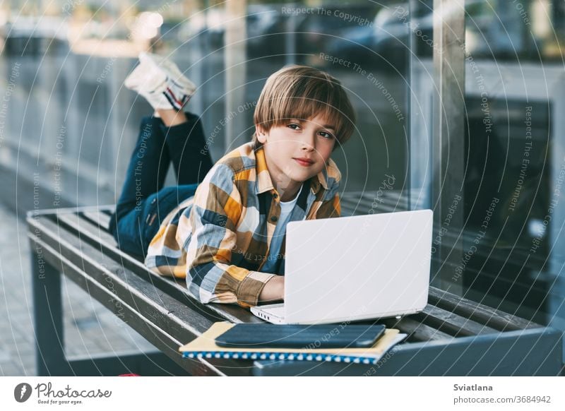 A smiling boy is lying on a bench and preparing for school lessons, next to a backpack. The boy is resting between classes. Social distance. Distance learning, education, modern technologies
