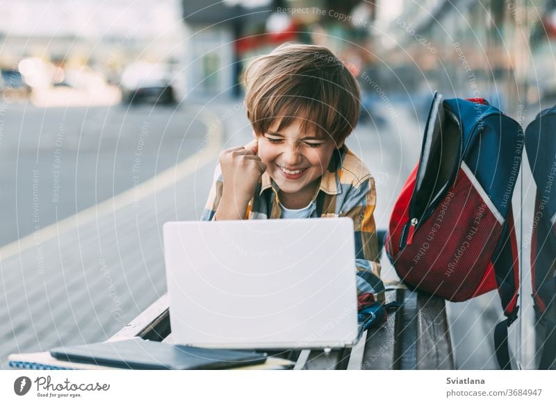 A cheerful boy is lying on a wooden bench and working on a laptop, next to a backpack. A student prepares for school lessons using the Internet. Social distance. Distance learning