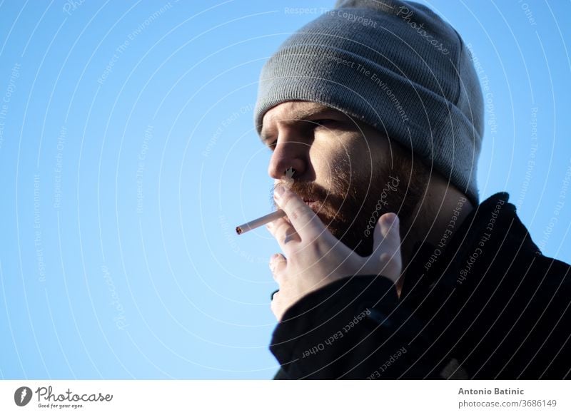 Caucasian bearded male with a beanie holding a cigarette, harshing sunlight with strong shadows.  Narrow shot with blue sky background addict addicted addiction