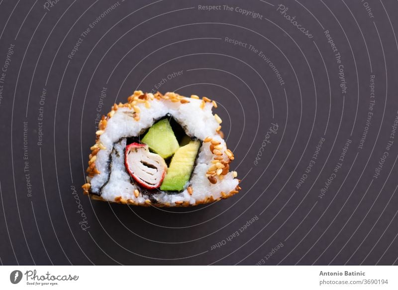 Closeup of a single piece of homemade not perfect sushi roll. Isolated on dark black background. Roll containing avocado, surimi, cucumber and fried sesame seeds