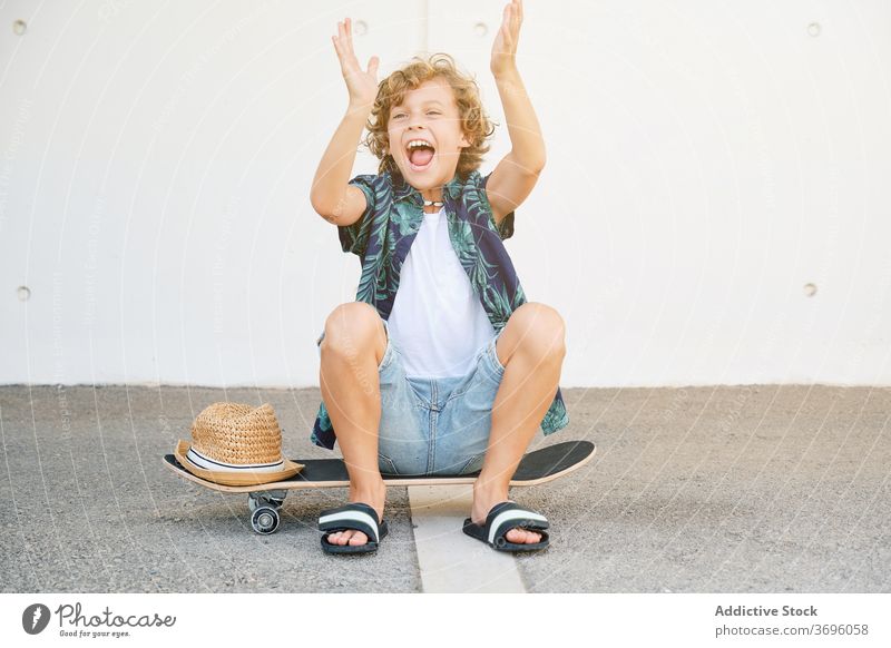 Child in summer clothes sitting on a skateboard gesturing joyfully dynamic extreme skill vitality skater skateboarder adolescence balance challenge children
