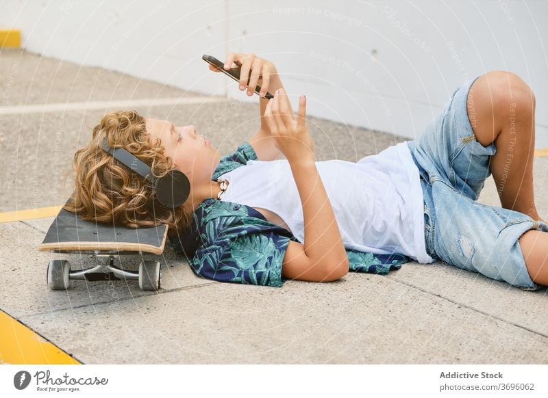 Boy lying on the floor with a skateboard listening to music and looking his mobile adolescence solitude singing trend generation teen weekend sound teenager