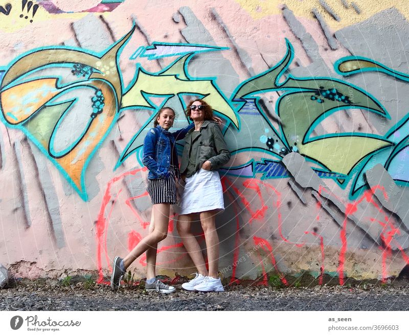 Two women in front of a wall with graffiti Graffiti sneakers Sunglasses lured Red-haired Exterior shot Day Human being Fashion green Khaki variegated Pattern