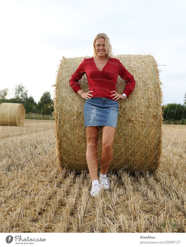 Young woman stands leaning against a bale of straw in the field and smiles Legs free time fun Joy jeans whole body Central perspective Looking into the camera