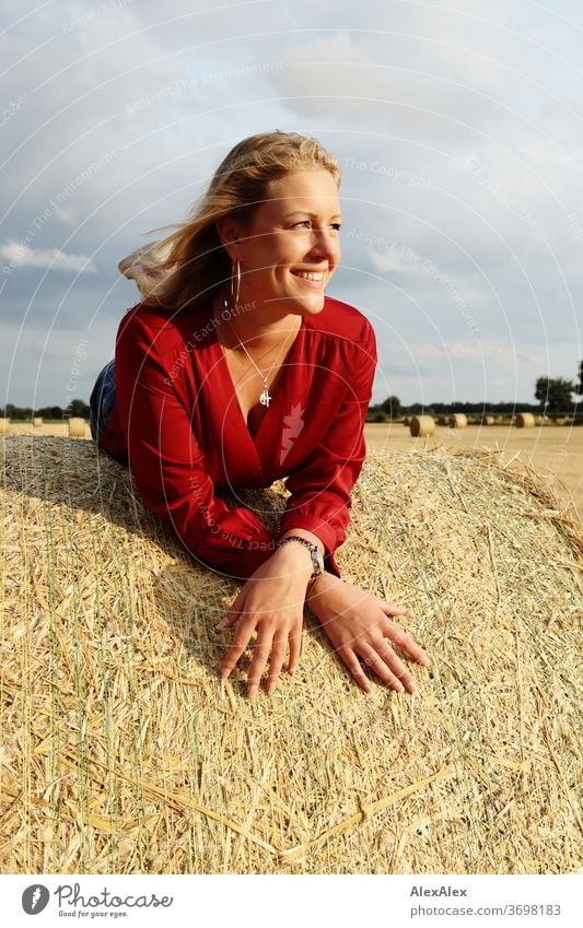 Young woman lies on a bale of straw in the field and smiles free time fun Joy jeans whole body Central perspective Looking into the camera