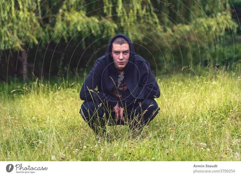 Portrait of a smiling, crouching man in a black hoodie portrait young smile happy lucky meadow nature summer look teenager looking male beautiful casual