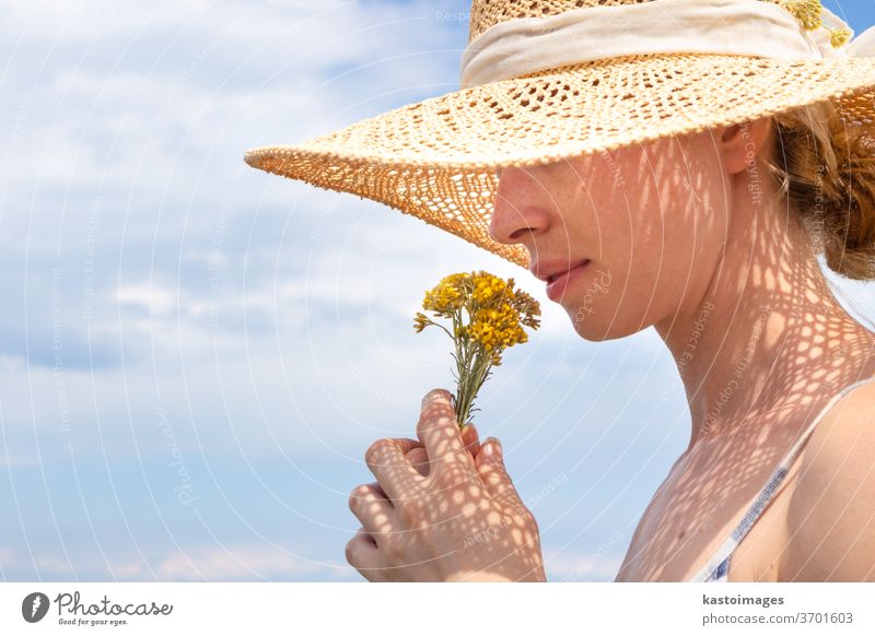 Portrait of young beautiful cheerful woman wearing straw sun hat, smelling small bouquet of yellow wild florets, against blue summer sky portrait flowers scent