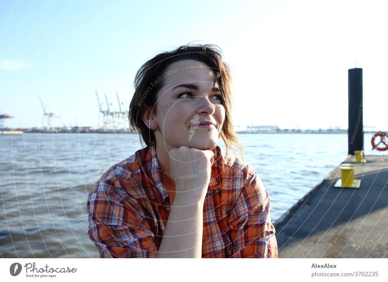 Young woman with freckles sitting on a pontoon in front of Hamburg harbour portrait Central perspective Looking brunette hair Copy Space right freckly Joy Model