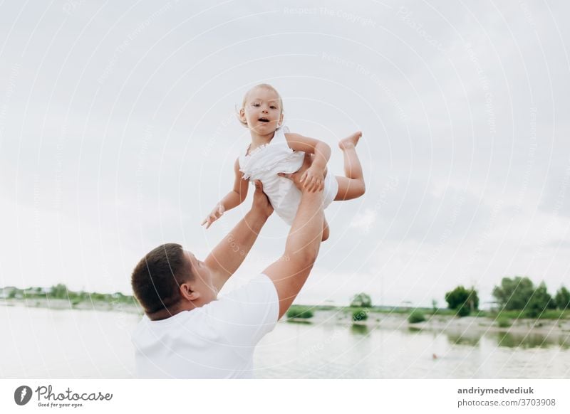 Father throws baby on pier on lake background, girl flies into the sky. Portrait father with child together. Daddy, little daughter outside. Family fishing holiday at pond.