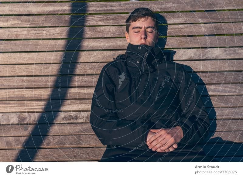 Young man sitting on a bench in the sun with his eyes closed young sunbath sunlight outdoors sleep relax leisure hands silent shadow wood portrait teenager
