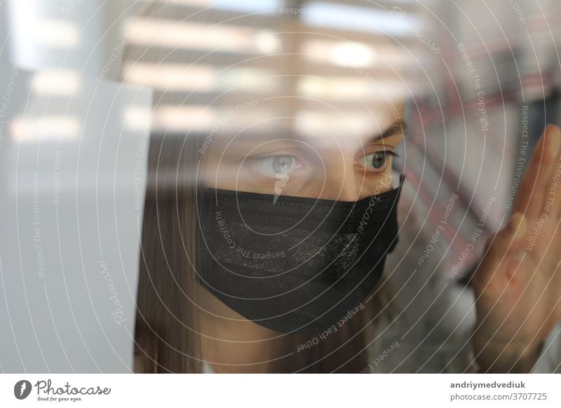young woman in a medical mask behind a windows glass at home. concept of quarantine of the coronavirus pandemic, covid 19. selective focus room epidemic illness